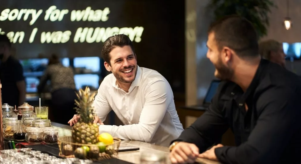 Two men leaning on a bar smiling at each other waiting to be served a drink.